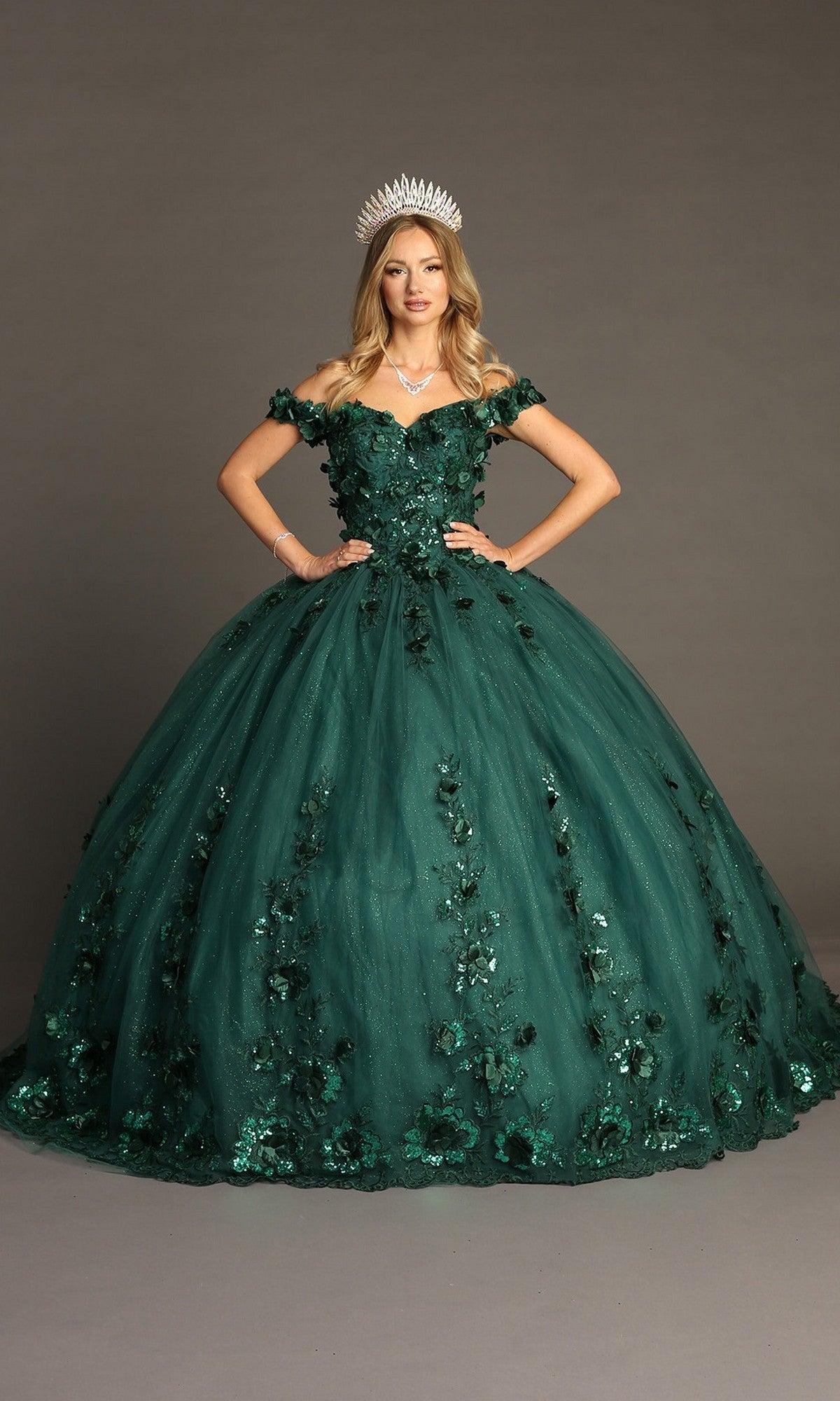 A woman in the Quinceanera Dress CHF3082 By Chicas—featuring a dark green hue, off-shoulder sleeves, 3D floral appliques, and a crown—stands against a plain background. The glitter tulle ballgown adds an enchanting touch to her regal look.
