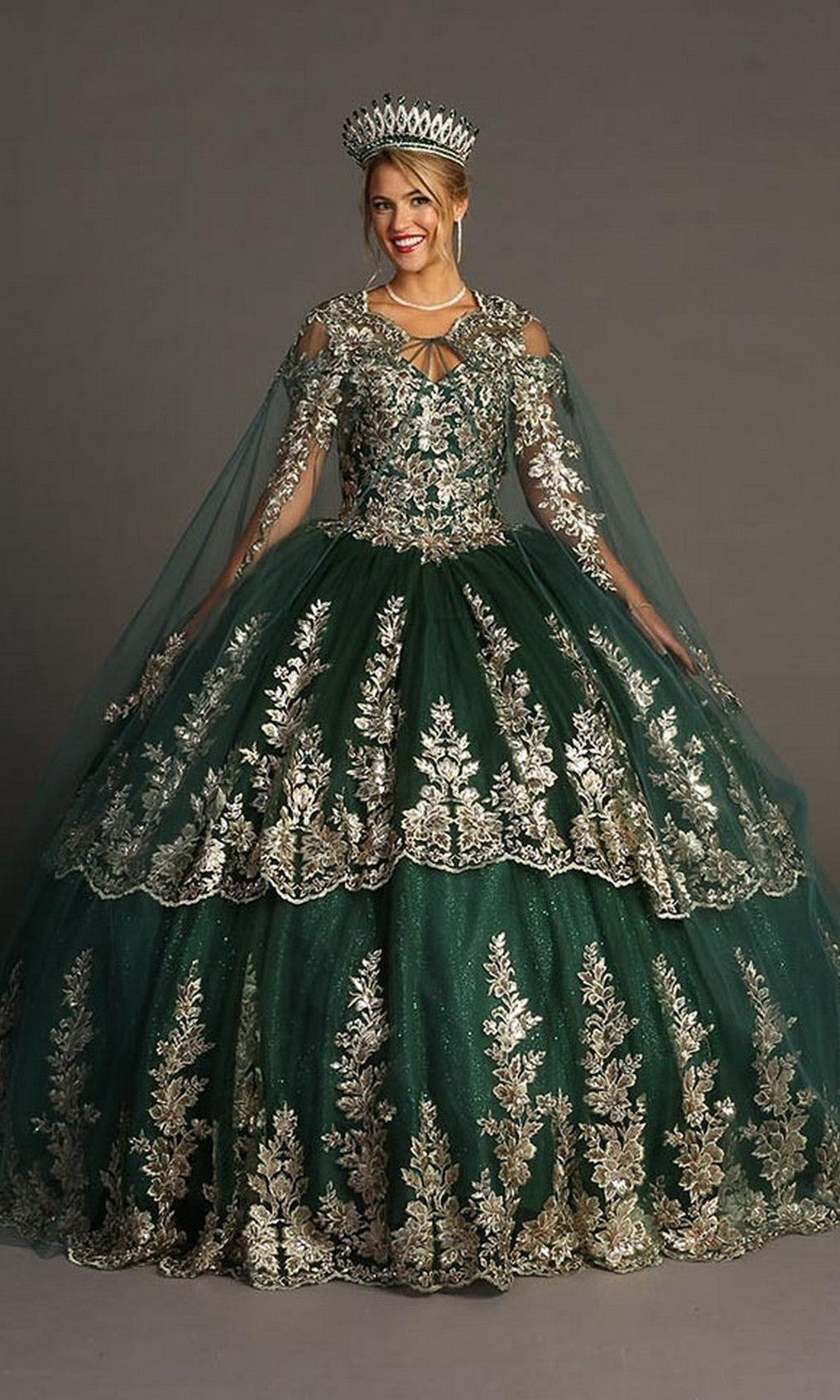 A woman stands against a plain background wearing the Quinceanera Dress CHF3083 By Chicas—an exquisite emerald green ballgown crafted from glitter tulle, detailed with gold embroidery, and paired with a matching cape and jeweled crown, evoking the regal elegance of a classic Quinceañera ensemble.
