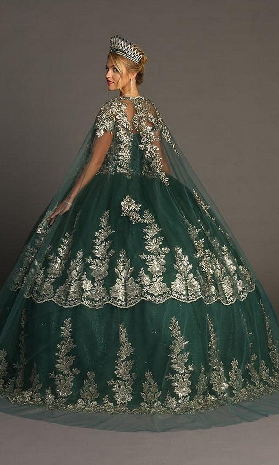 A woman stands gracefully against a plain backdrop, elegantly crowned and dressed in the enchanting Quinceanera Dress CHF3083 By Chicas—a large green ballgown adorned with glitter tulle and silver floral embroidery—capturing the regal essence of a traditional Quinceañera celebration.