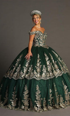 A woman wearing the Quinceanera Dress CHF3083 by Chicas—an ornate green gown with gold embroidery and a sparkling glitter tulle ballgown skirt—stands against a plain gray background, smiling as she completes her look with a matching crown.