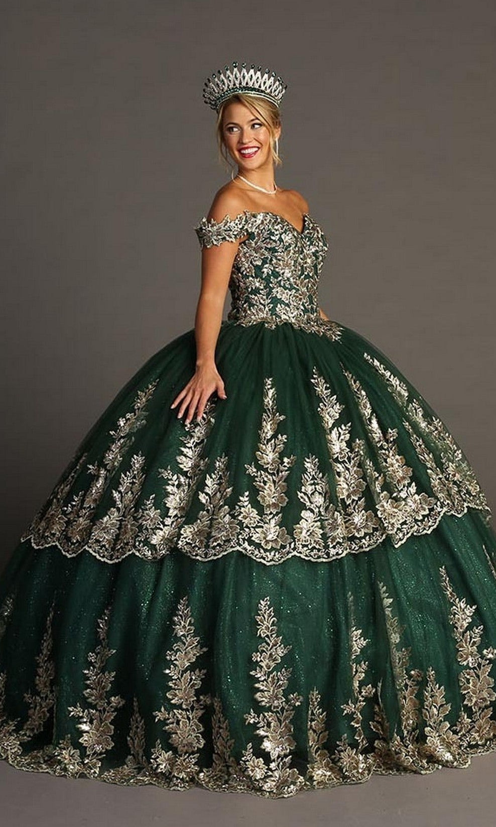 A woman stands and smiles against a plain gray background, wearing the Quinceanera Dress CHF3083 By Chicas—an off-the-shoulder, sweetheart neckline dark green gown adorned with gold embroidery and paired with a matching crown.