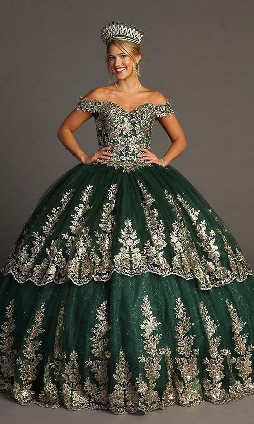 A woman stands smiling against a plain background, wearing the Quinceanera Dress CHF3083 By Chicas in green. The off-shoulder gown features gold embroidery and matching crown, with layers of glitter tulle that enhance the elegance of her radiant appearance.