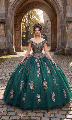 A woman wearing the Quinceanera Dress CHF3084 by Chicas—a stunning green glitter tulle ballgown adorned with gold floral embroidery—stands on a cobblestone path in front of a stone archway and historic building.