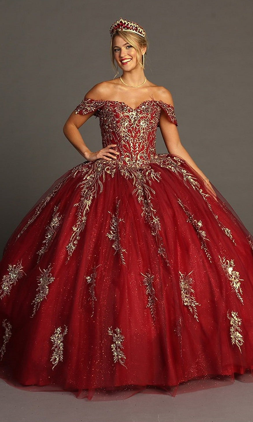 A woman wearing the Quinceanera Dress CHF3084 By Chicas—a stunning red off-the-shoulder ball gown embellished with gold floral embroidery and paired with a matching tiara—stands against a plain gray background.
