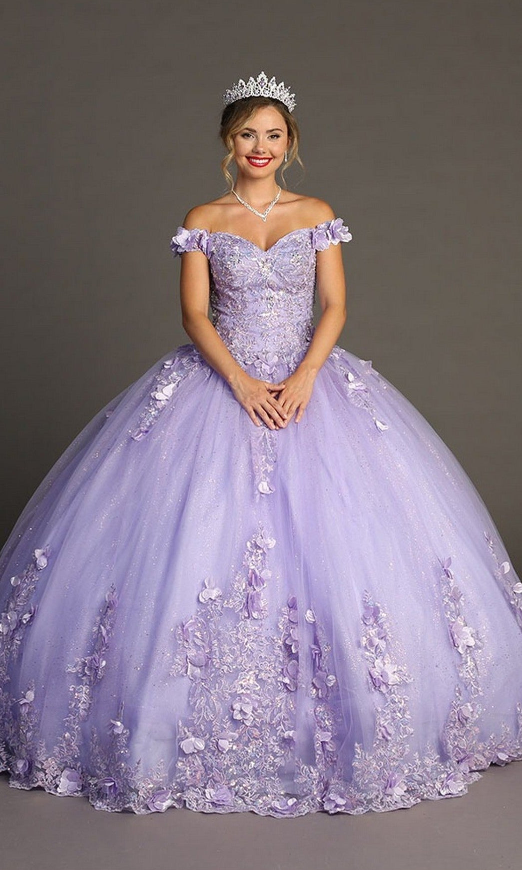 A woman wears the Quinceanera Dress CHF3085 By Chicas—a glitter tulle ballgown adorned with delicate floral appliques—along with a tiara and necklace, standing against a plain gray background.