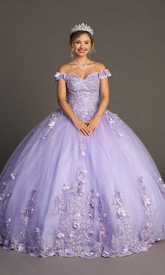 A woman wears the Quinceanera Dress CHF3085 By Chicas—a glitter tulle ballgown adorned with delicate floral appliques—along with a tiara and necklace, standing against a plain gray background.