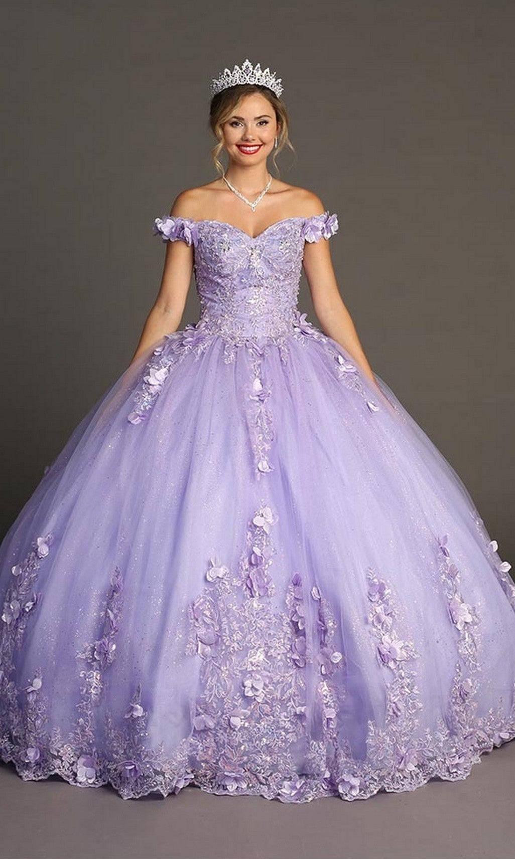 A woman wearing the Quinceanera Dress CHF3085 By Chicas in lilac, featuring off-shoulder styling and floral appliqués, paired with a tiara, necklace, and red lipstick, stands against a plain gray background.