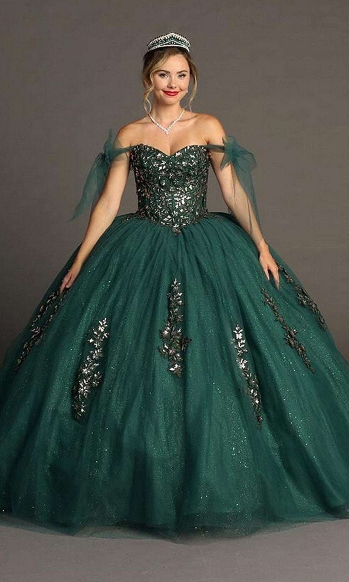 A woman wearing the Quinceanera Dress CHF3086 By Chicas in green, adorned with silver embellishments and featuring off-the-shoulder sleeves along with a voluminous ballgown skirt, poses gracefully against a plain gray backdrop, her look completed with a sparkling tiara.