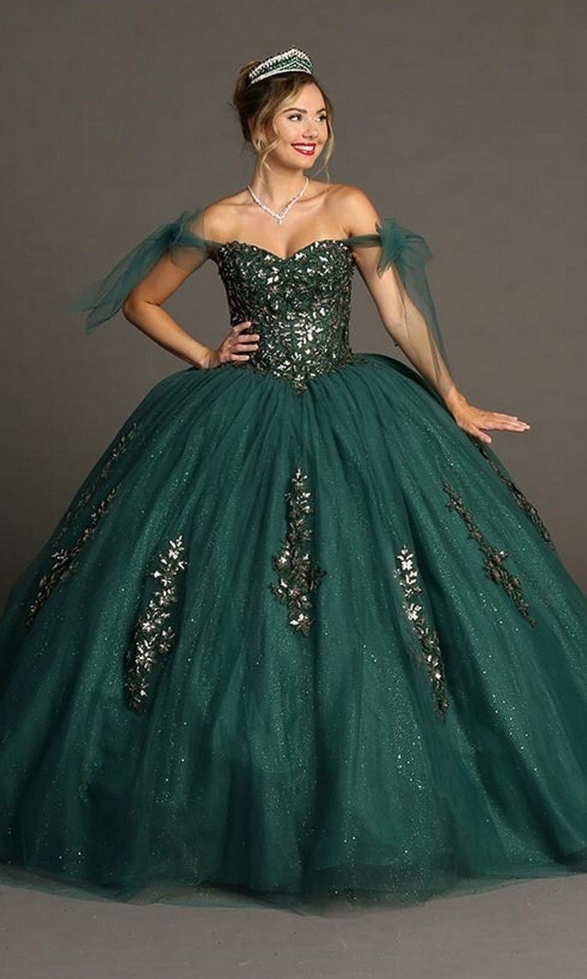 A woman wearing the Quinceanera Dress CHF3086 By Chicas, a dark green off-the-shoulder gown adorned with silver embellishments, paired with a tiara and necklace, poses against a plain background.
