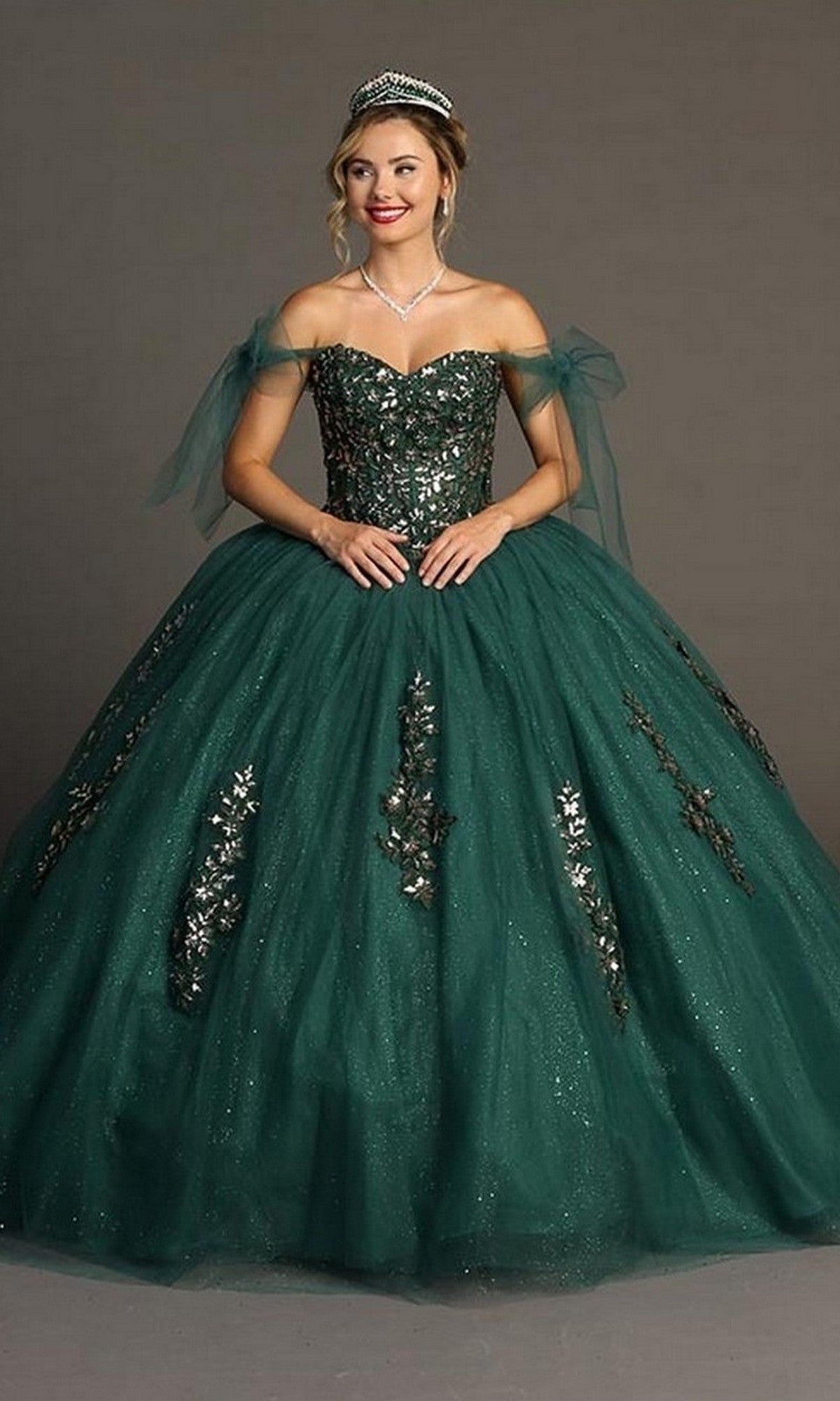 A woman stands against a plain background wearing the Quinceanera Dress CHF3086 By Chicas, a dark green off-the-shoulder gown adorned with silver embellishments and paired with a tiara, beautifully showcasing the elegance of its traditional voluminous ballgown skirt.