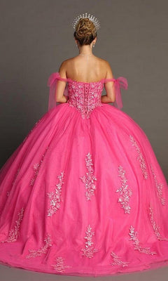 A woman wearing the Quinceanera Dress CHF3086 by Chicas—a bright pink off-the-shoulder ballgown skirt with delicate floral embroidery and a tiara—stands facing away against a plain background.