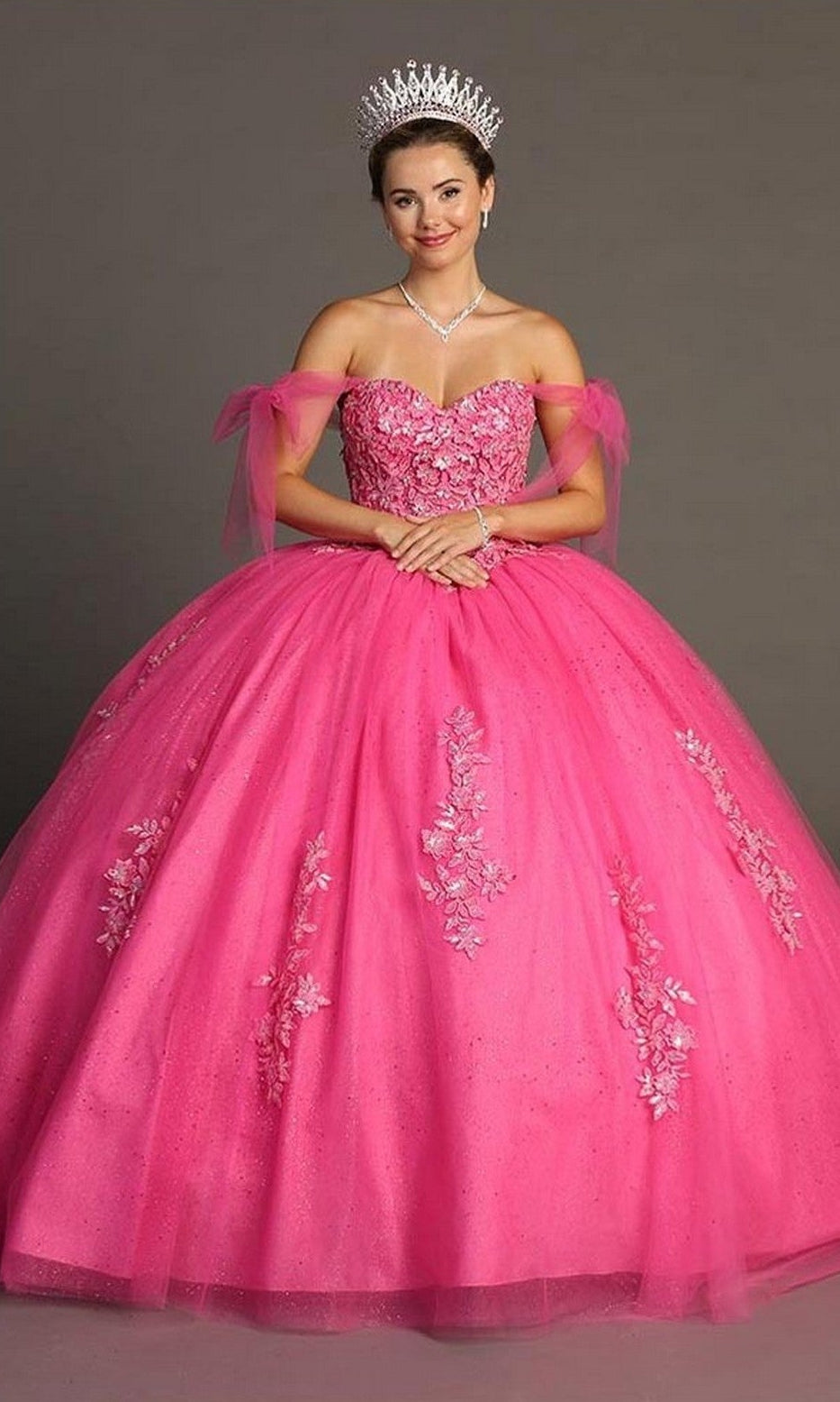Wearing the Quinceanera Dress CHF3086 By Chicas, a woman stands against a plain background with her hands folded. The dress’s vibrant pink color, floral details, jeweled tiara, off-the-shoulder bodice, and voluminous ballgown skirt create an elegant and sophisticated look.