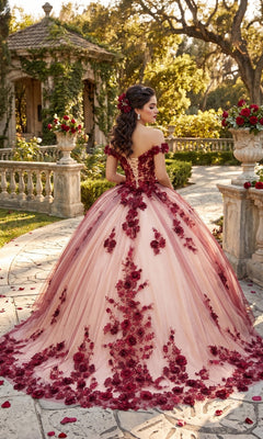 Wearing the Quinceanera Dress CHF3088 By Chicas, a woman stands outdoors on a stone path, surrounded by greenery and scattered rose petals. The off-the-shoulder ballgown features pink tulle adorned with red floral appliqué.