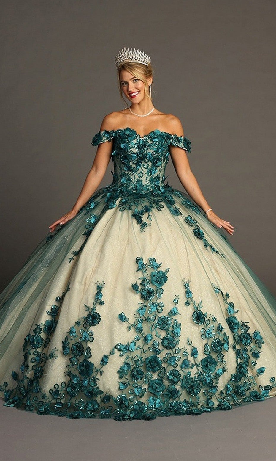A woman stands against a plain gray background wearing the Quinceanera Dress CHF3088 By Chicas in green/nude, an off-the-shoulder gown adorned with teal floral embroidery and featuring a voluminous tulle ballgown skirt. She accessorizes with a pearl necklace and a silver crown.