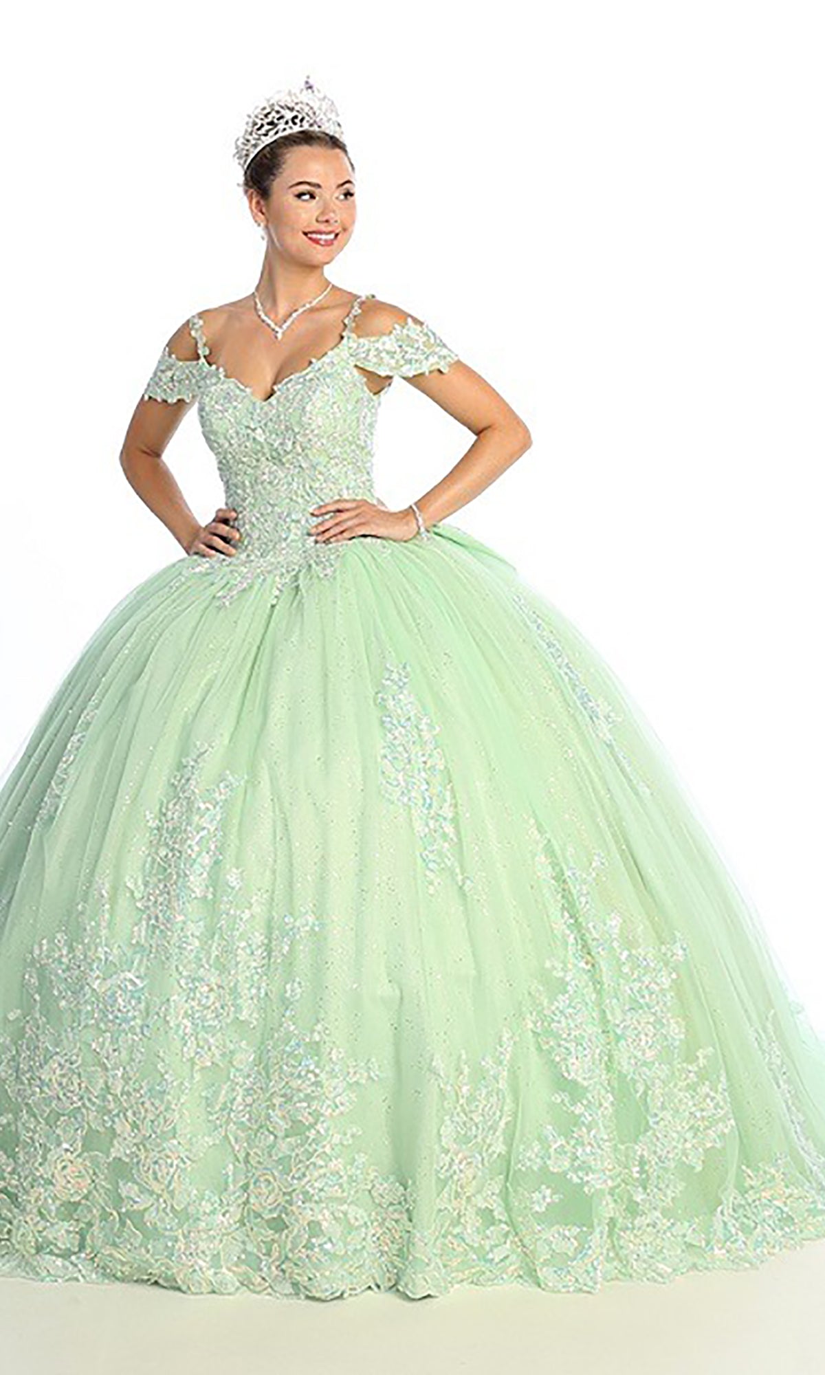 A woman wearing the Quinceanera Dress CHF3089 By Chicas in sage, featuring a cold shoulder design, floral lace corset bodice, and delicate lace details, paired with a tiara and jewelry, stands against a white background.