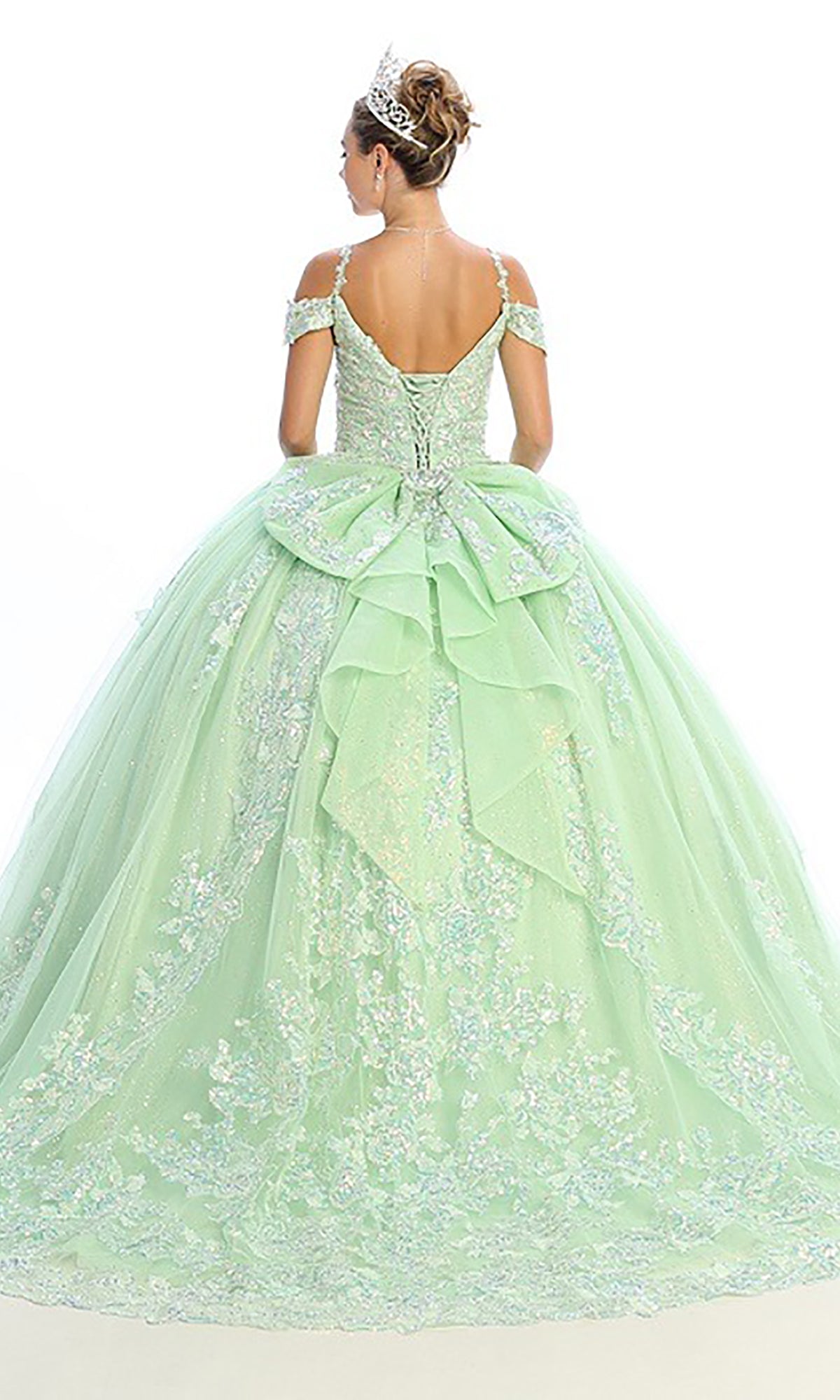 A woman faces away, wearing the Quinceanera Dress CHF3089 By Chicas—a light green, off-shoulder ball gown featuring a glitter-tulle skirt, corset bodice adorned with floral lace details, and a large bow—paired with a tiara.