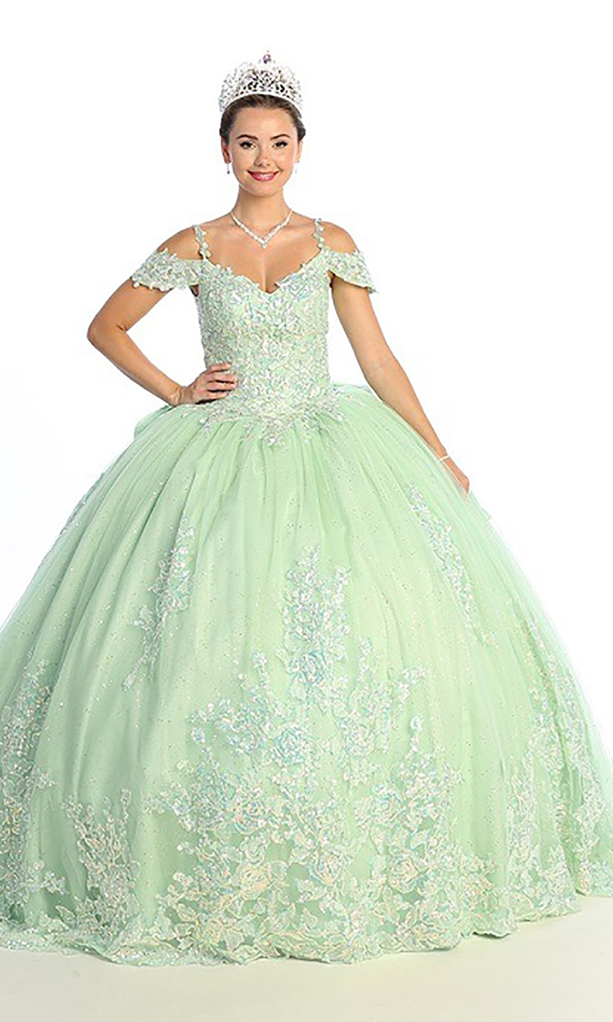 A young woman wearing the Quinceanera Dress CHF3089 By Chicas, a light green glitter-tulle ballgown featuring off-the-shoulder sleeves and a floral lace corset bodice, accessorized with a tiara and necklace, stands against a plain white background.