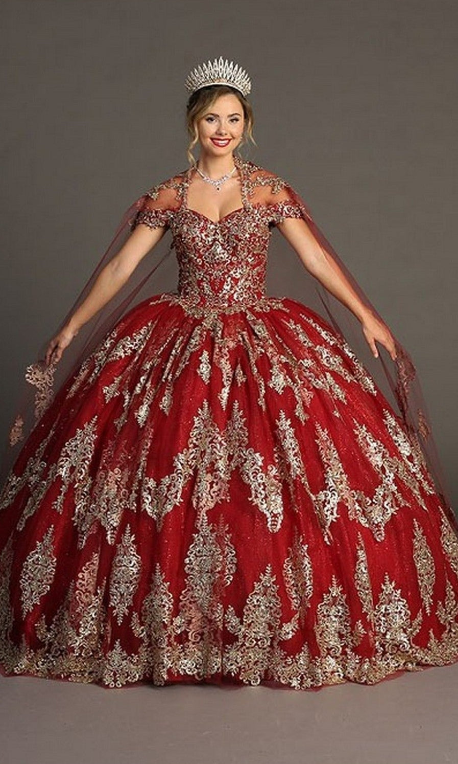 A woman dressed in the Quinceanera Dress CHF3090 By Chicas in burgundy and gold with glitter tulle and gold embroidery, accessorized with a silver tiara and jewelry, stands against a plain background.