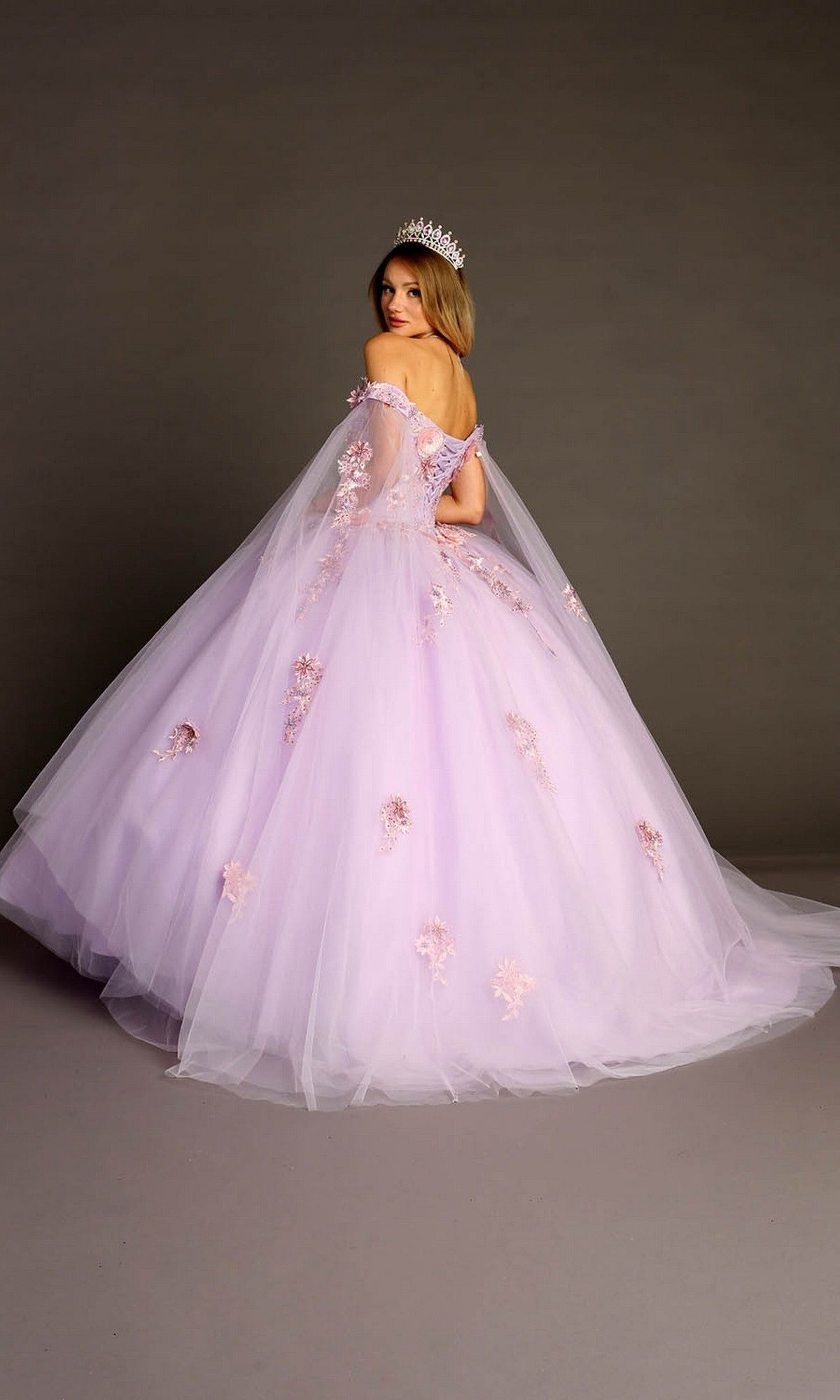 A woman wears the Quinceanera Dress CHF3095 By Chicas—a lilac ballgown embellished with floral details and paired with a tiara—standing on a gray studio backdrop, turned slightly away from the camera.
