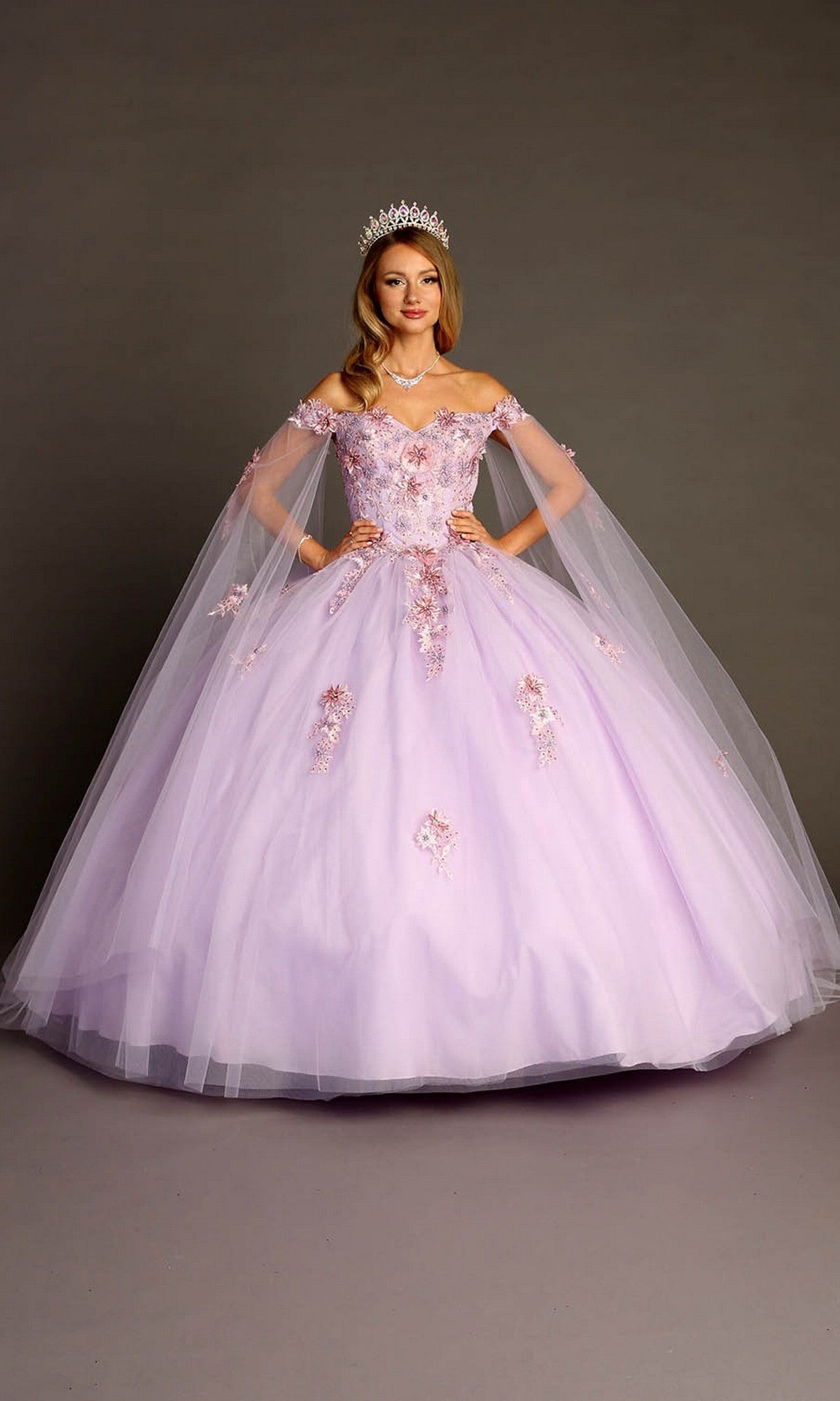 A woman stands against a plain background wearing the Quinceanera Dress CHF3095 by Chicas—a lilac ballgown adorned with floral appliques and featuring delicate tulle sleeves—paired with a tiara.