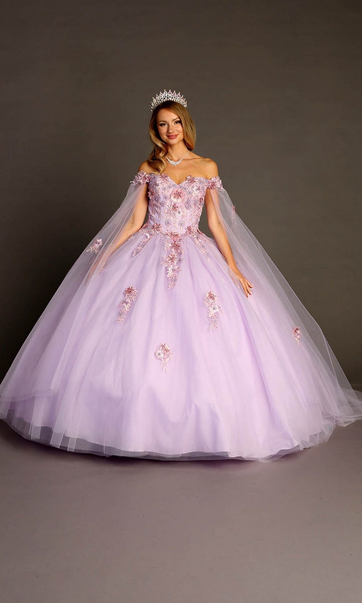A person wearing the Quinceanera Dress CHF3095 by Chicas—with delicate floral appliques, off-the-shoulder sleeves, a voluminous lilac tulle skirt, and a sparkling tiara—stands gracefully in front of a plain grey background.
