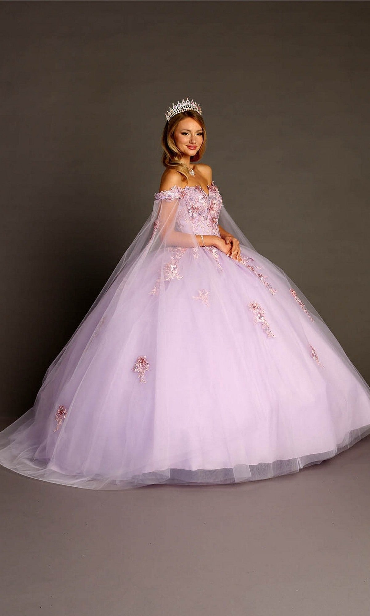 A woman wearing a sparkling tiara and the Quinceanera Dress CHF3095 By Chicas in lilac, beautifully adorned with intricate floral appliques and featuring an off-the-shoulder design, stands against a plain studio background.