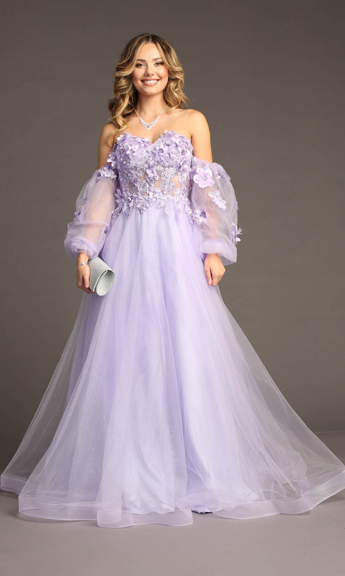 A woman stands against a plain background wearing the Chicas CHF3096 Long Sleeve Long Prom Ball Gown—a light purple off-the-shoulder dress with floral appliques and sheer sleeves—holding a silver clutch.