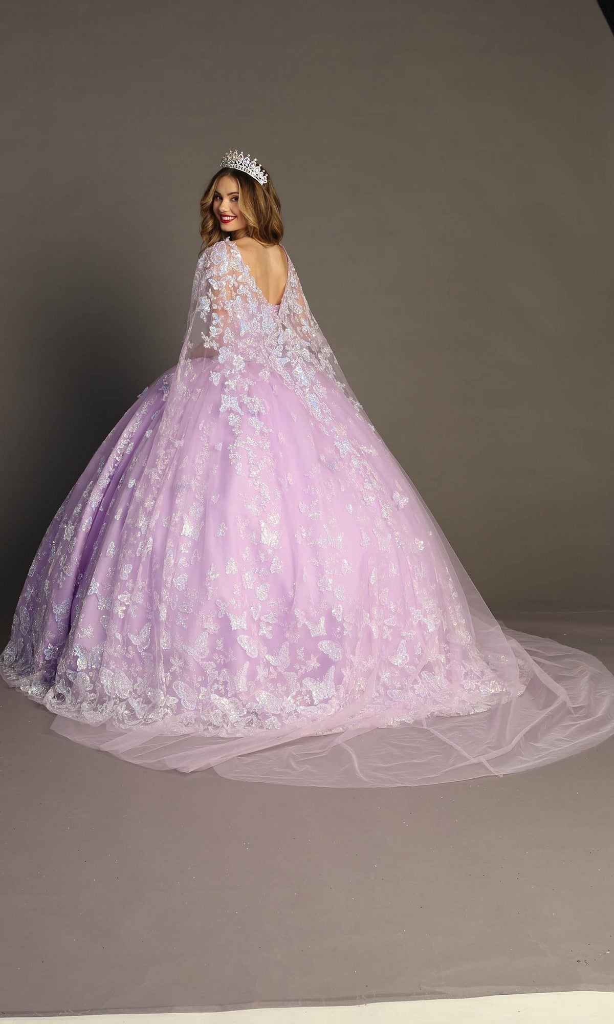A woman in the Quinceanera Dress CHF3097 By Chicas—featuring a lavender hue, floral details, delicate butterfly appliques, and a sparkling glitter tulle skirt—stands against a gray backdrop, facing away but glancing back over her shoulder with a smile as her tiara gleams softly.