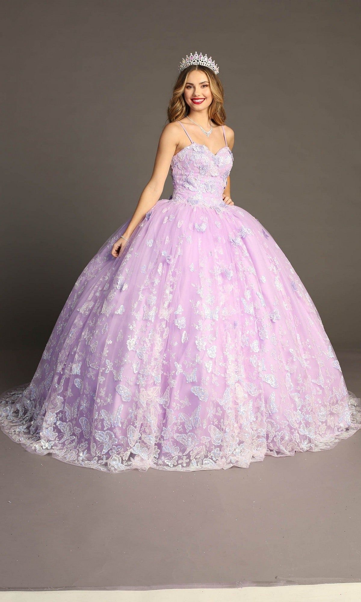 A woman wearing the Quinceanera Dress CHF3097 By Chicas—a lavender gown adorned with floral embellishments, butterfly appliques, and a glitter tulle skirt—stands against a plain background, smiling and holding the skirt of her gown while wearing a tiara.