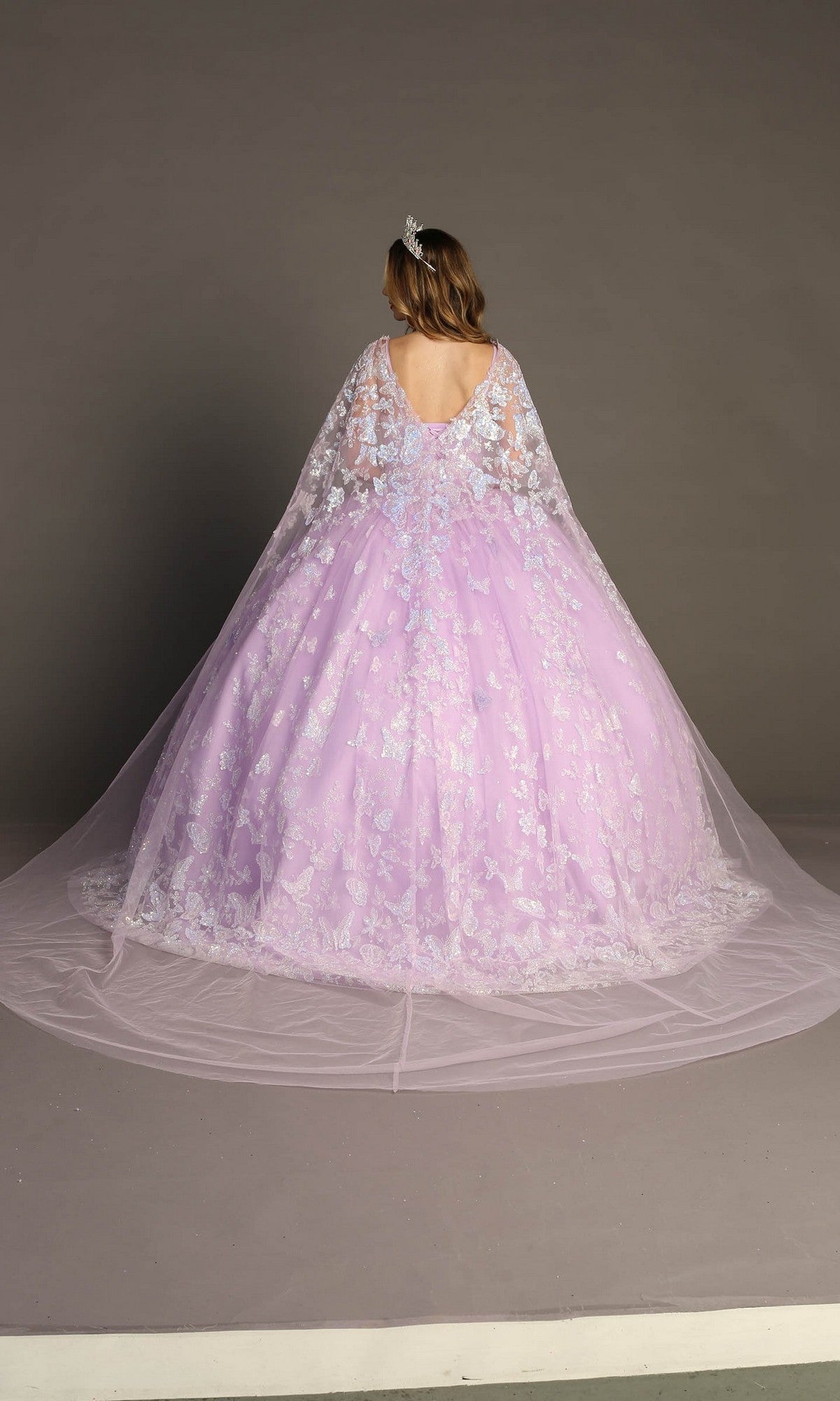 Person standing with their back to the camera, wearing the Quinceanera Dress CHF3097 By Chicas—a voluminous lavender gown featuring lace details and a sheer cape embellished with delicate butterfly appliques—set against a plain gray background.