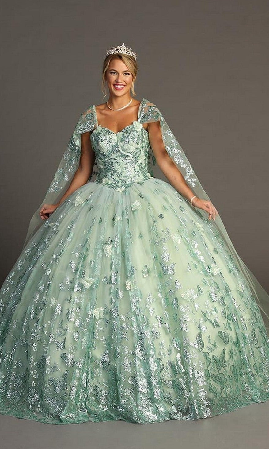 A woman wearing the Quinceanera Dress CHF3097 By Chicas in sage—a sequin-embellished ball gown with sheer cape sleeves and glitter tulle accents—paired with a tiara and pearl necklace, stands against a plain gray background.