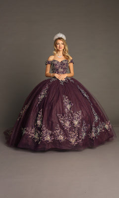 A woman wearing a sparkling crown and an ornate, off-the-shoulder Quinceanera Dress CHF3099 By Chicas in purple, featuring a flowing ballgown skirt and delicate floral embroidery, stands against a plain gray background.