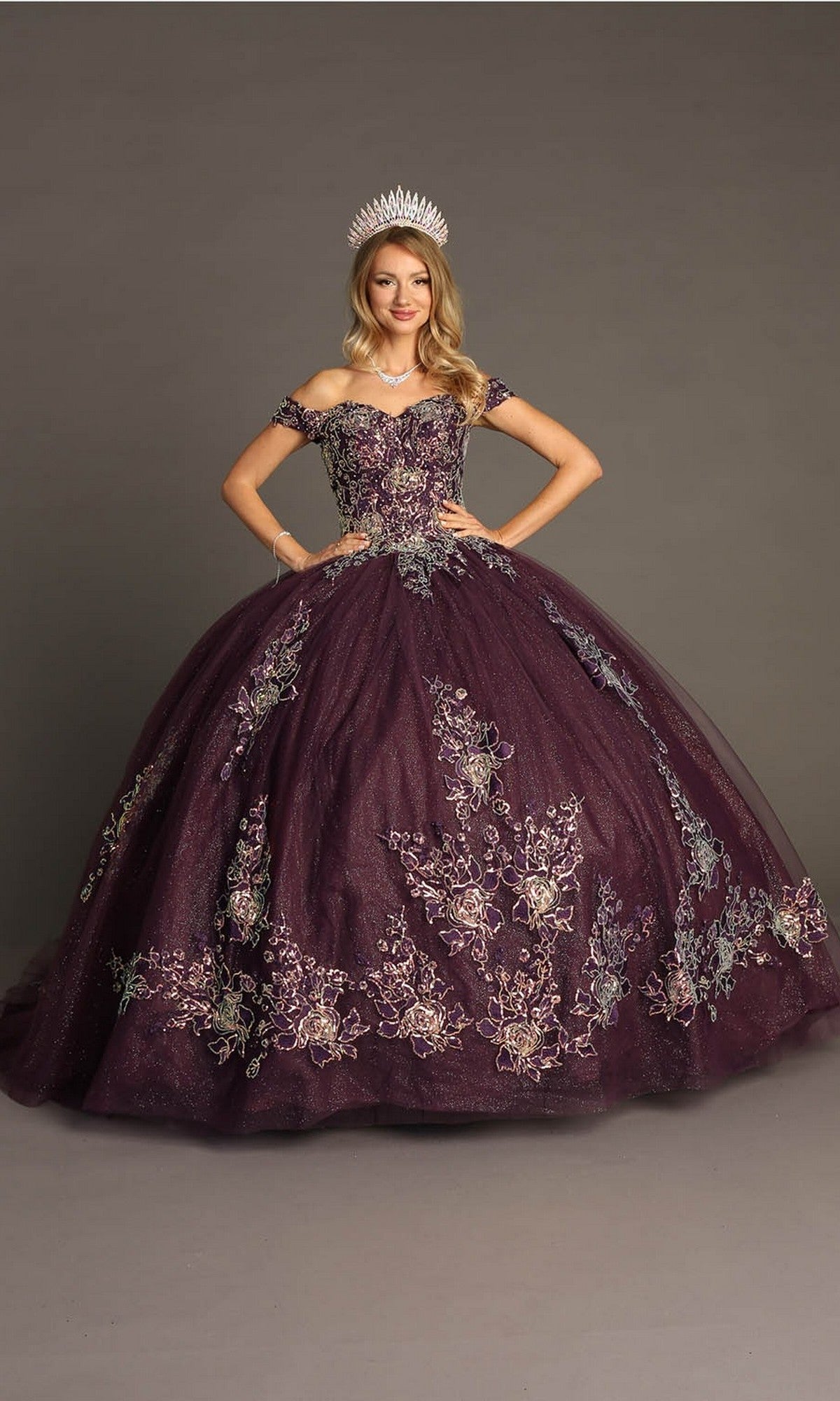 A woman wearing the Quinceanera Dress CHF3099 By Chicas—a dark purple off-the-shoulder ballgown skirt with floral embroidery—paired with a silver tiara, stands against a plain background.