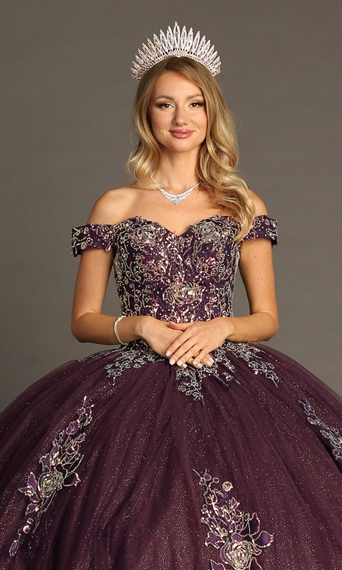 A woman wearing the Quinceanera Dress CHF3099 By Chicas—a jeweled crown adorning her head, this off-the-shoulder purple gown features intricate floral embroidery and a voluminous ballgown skirt—as she poses against a neutral background.