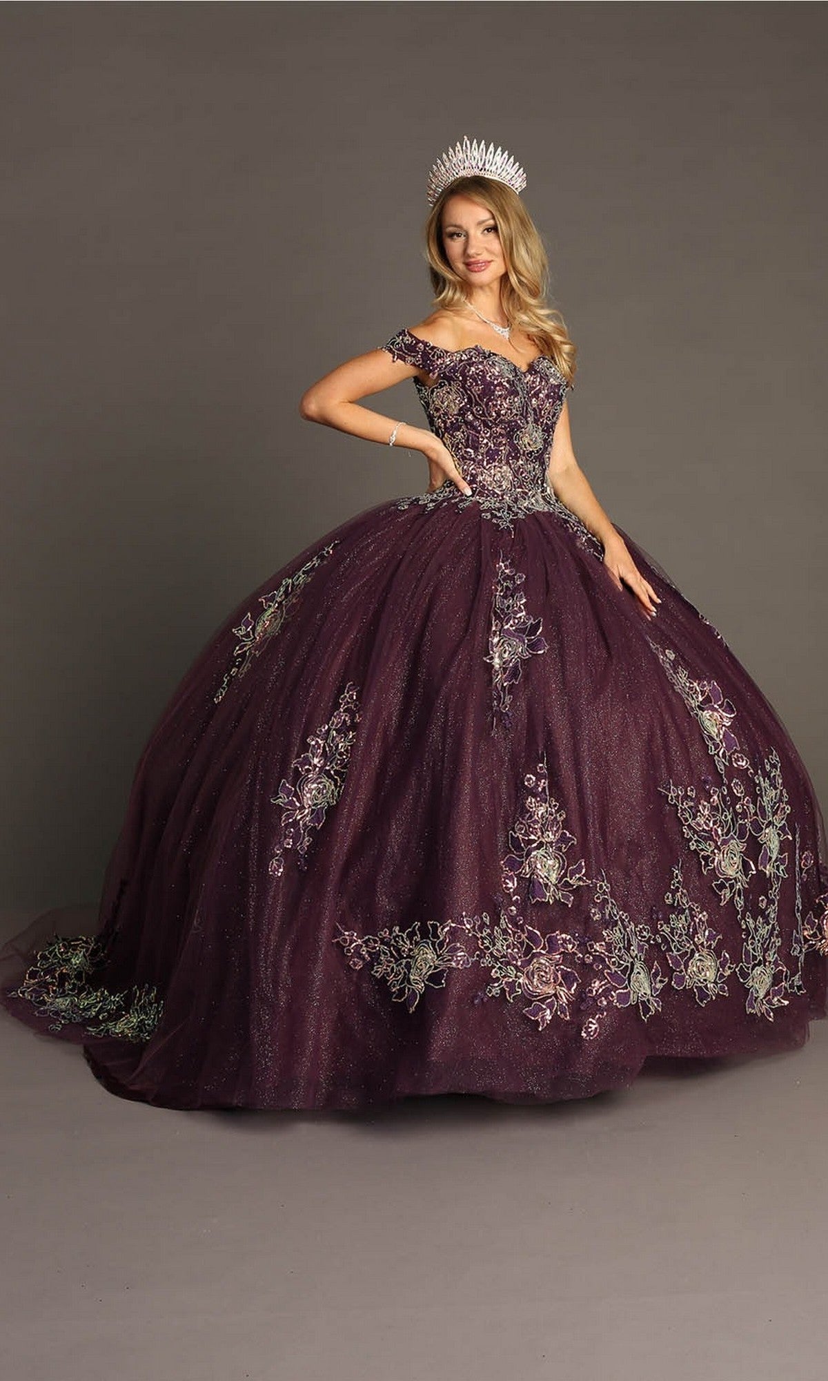 A woman wearing the Quinceanera Dress CHF3099 by Chicas, a purple off-the-shoulder ballgown skirt with floral embroidery, paired with a silver tiara and standing against a plain gray background.