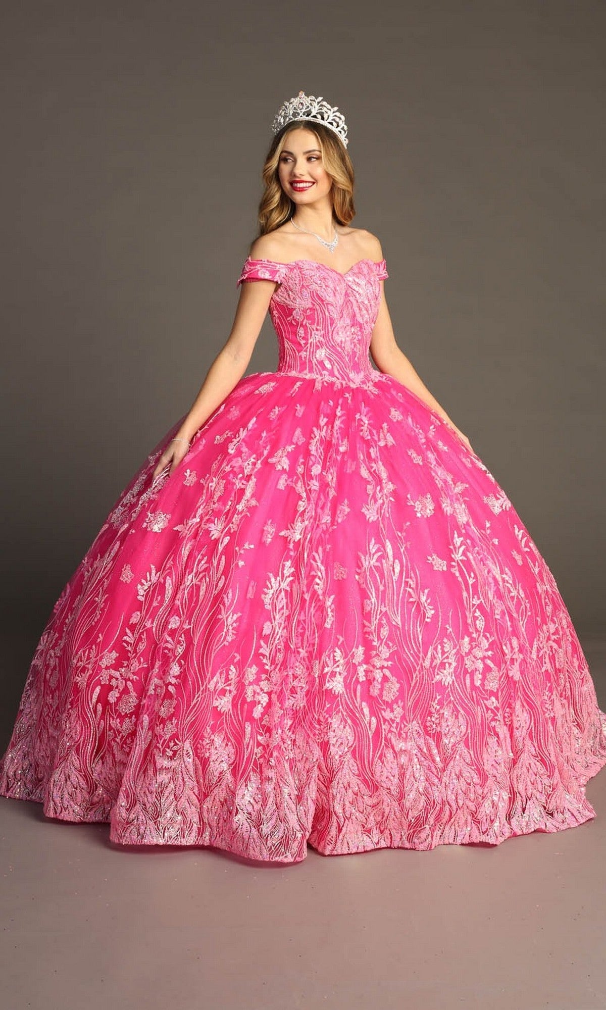 Wearing a sparkling tiara, a woman poses against a plain background in the Quinceanera Dress CHF3100 By Chicas—an off-the-shoulder, bright pink gown adorned with elegant white floral patterns.