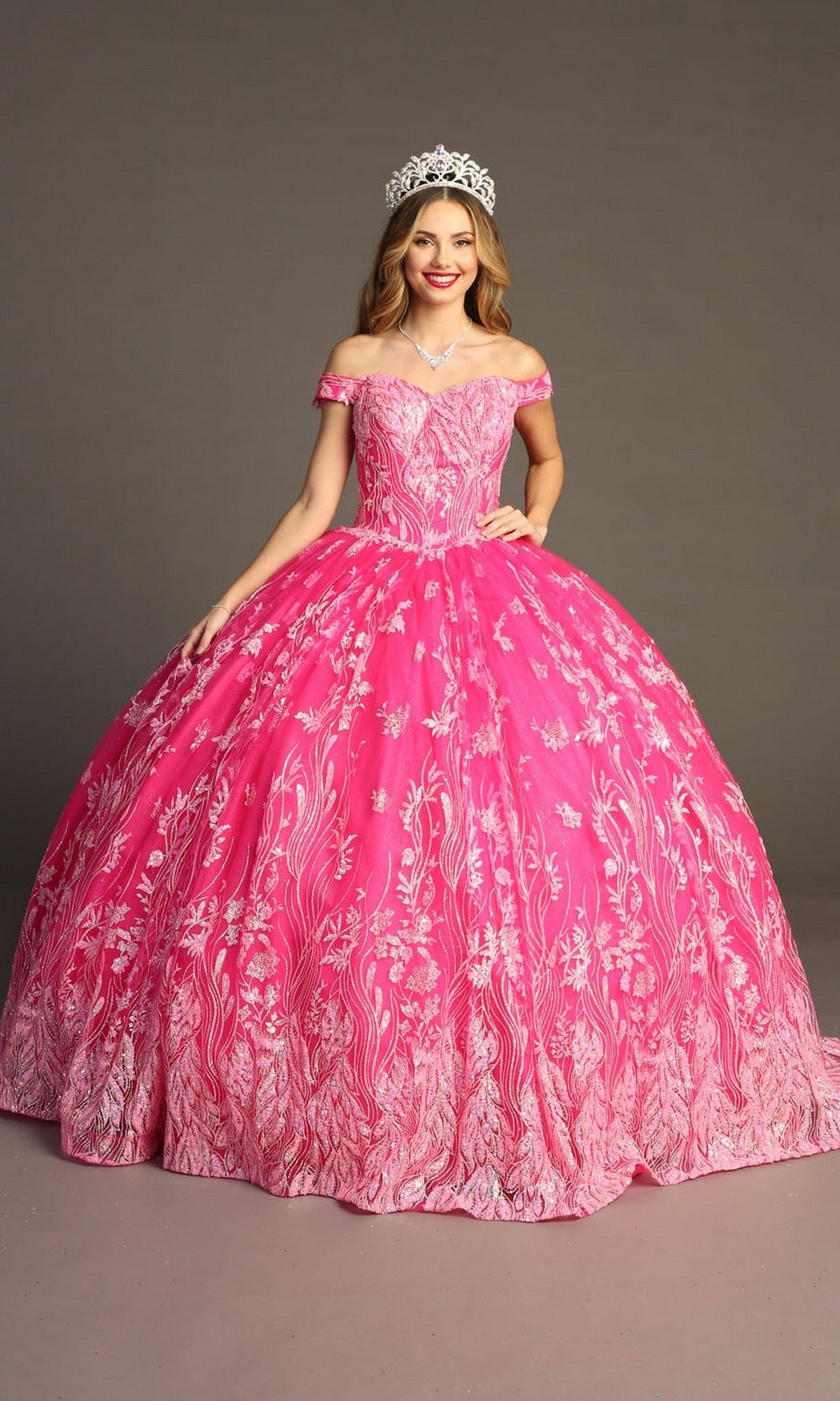A woman wearing the Quinceanera Dress CHF3100 By Chicas—an off-the-shoulder pink tulle ballgown with white floral patterns—stands against a plain backdrop, smiling and accessorized with a jeweled tiara.