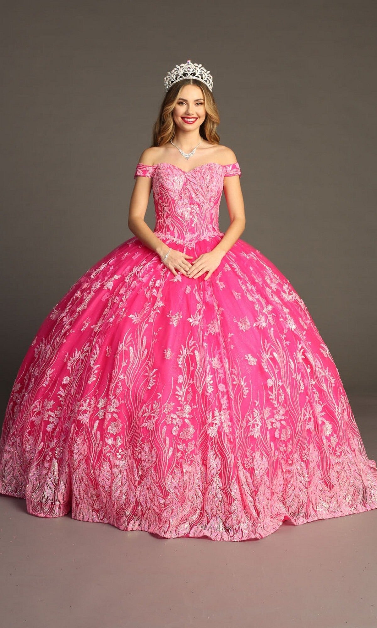 A young woman stands against a plain backdrop, smiling at the camera in the Quinceanera Dress CHF3100 By Chicas—a bright pink, off-the-shoulder gown featuring delicate floral patterns and a flowing tulle ballgown skirt. She completes her look with a jeweled tiara and necklace.