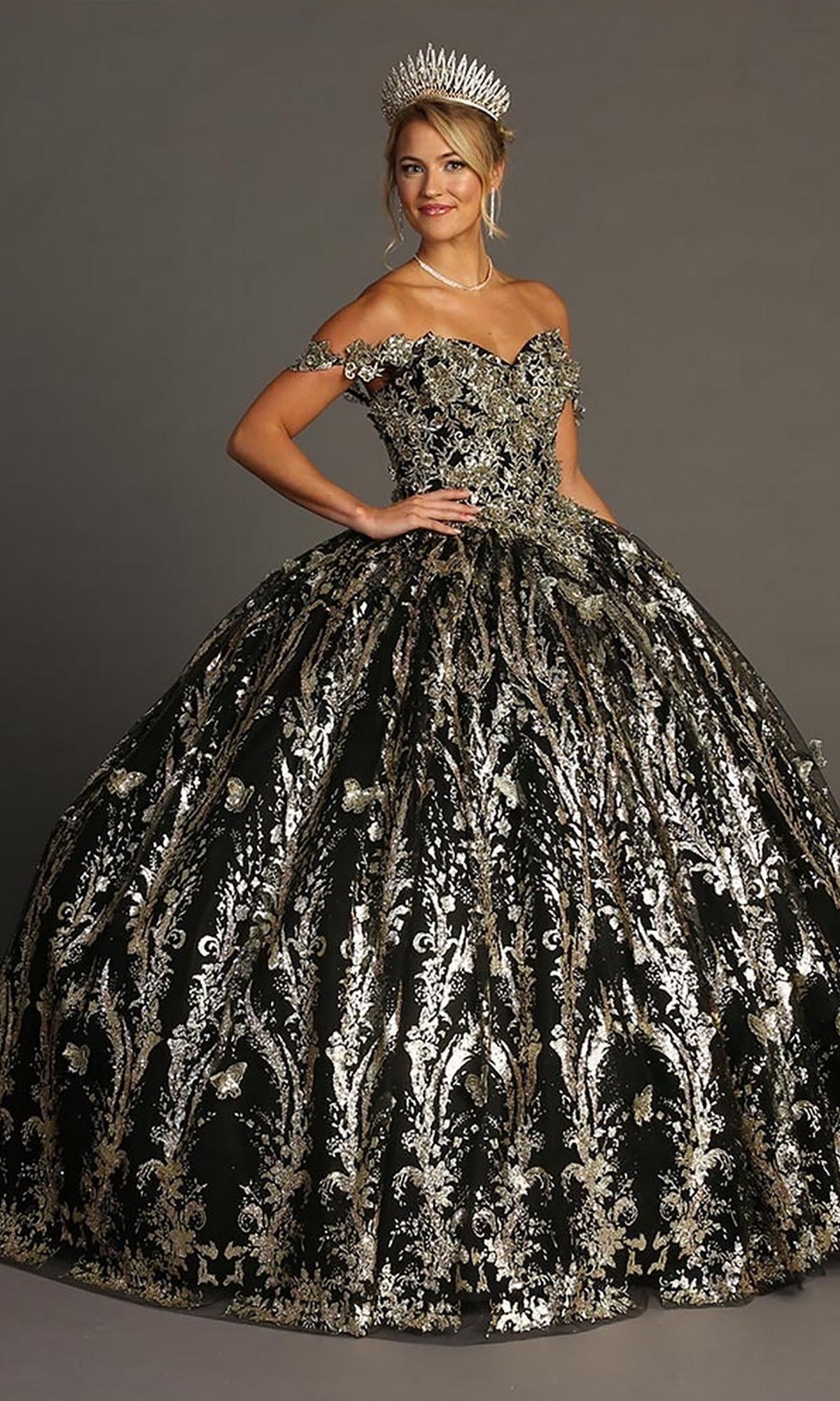 A woman wearing the Quinceanera Dress CHF3101 By Chicas in black and gold, featuring a voluminous tulle ballgown skirt and gold embellishments, with a tiara, poses against a plain gray background.