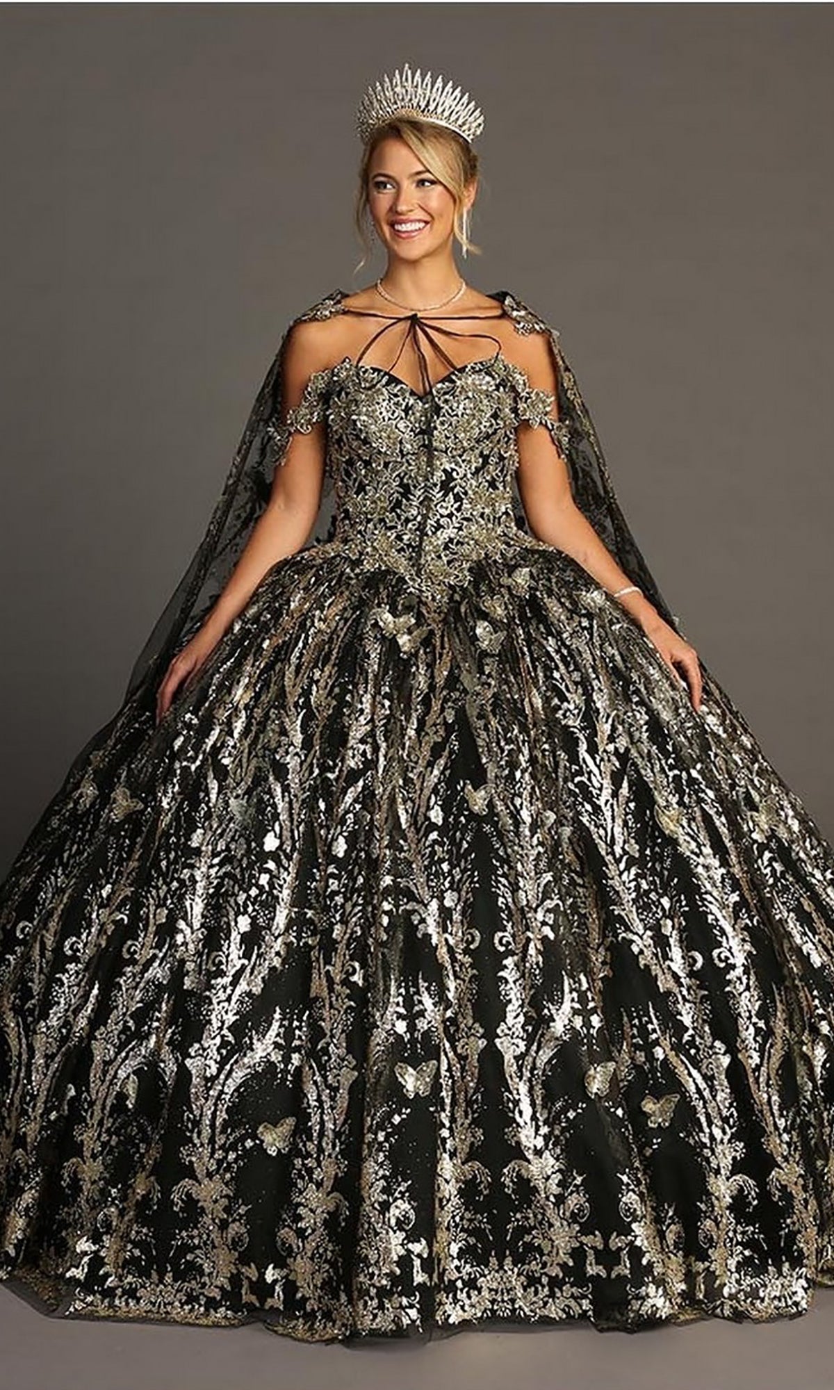 A woman wearing the Quinceanera Dress CHF3101 By Chicas—an ornate black and gold off-the-shoulder gown with a full tulle ballgown skirt, detailed with intricate patterns, metallic-silver embellishments, and paired with a silver tiara—stands against a plain background.