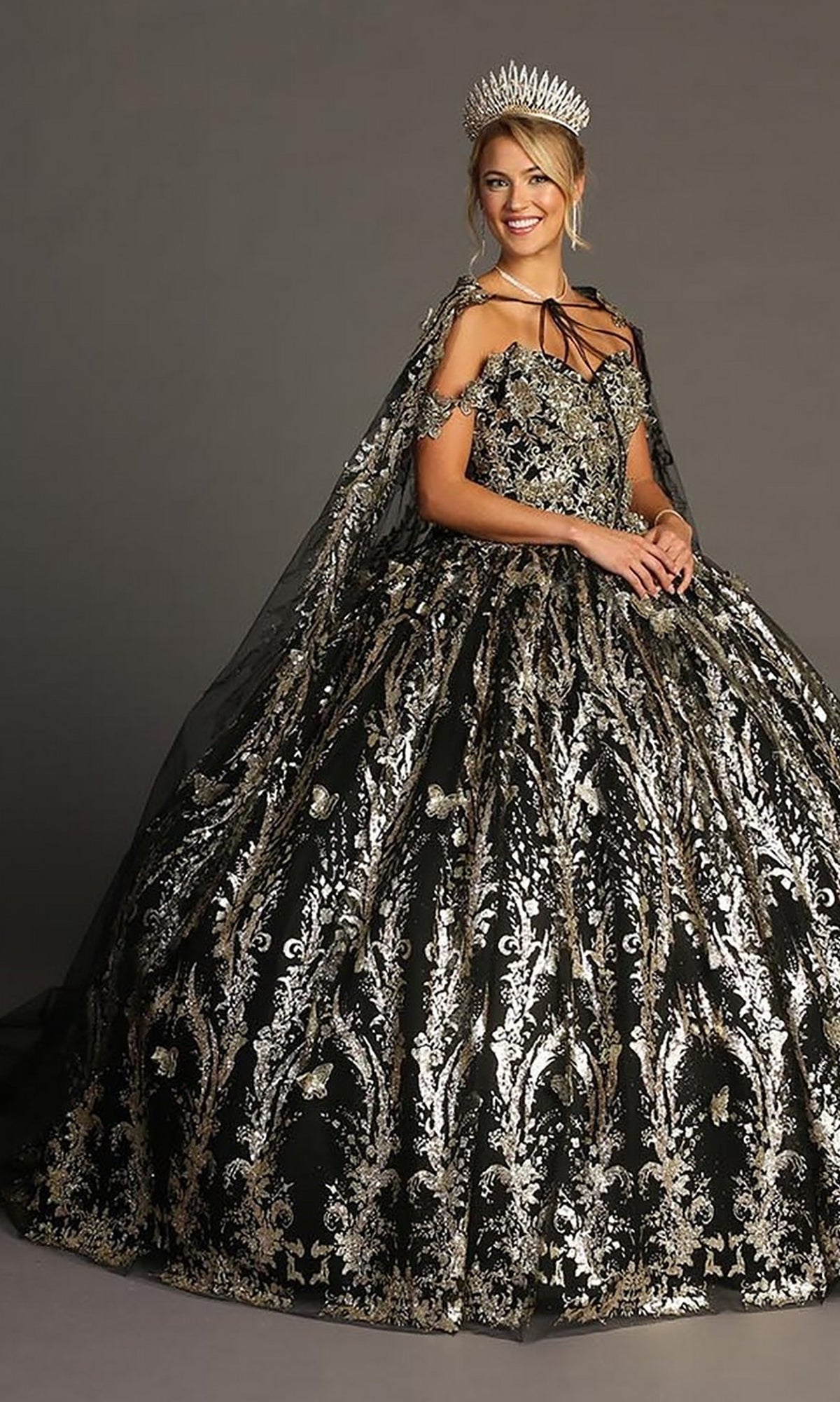 A woman wearing the Quinceanera Dress CHF3101 by Chicas—an ornate black ball gown adorned with metallic-silver embellishments and a dramatic tulle skirt—paired with a matching cape and a large silver crown, stands against a plain background.