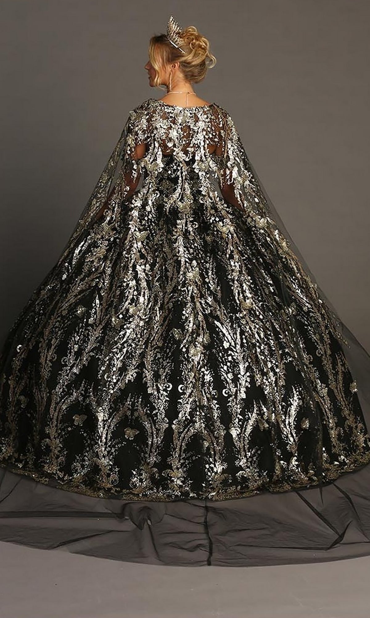 A woman in a silver tiara wears the Quinceanera Dress CHF3101 By Chicas—an off-the-shoulder black ball gown adorned with silver floral embroidery and metallic-silver embellishments, featuring a sheer, matching cape and a dramatic tulle skirt. She stands against a gray background.