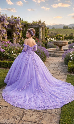 Wearing the Quinceanera Dress CHF3102 By Chicas, a woman stands on a stone path in a garden, her back turned toward blooming flowers and a fountain. The off-the-shoulder gown features 3D floral appliques, and the tulle ballgown skirt trails gracefully behind her.