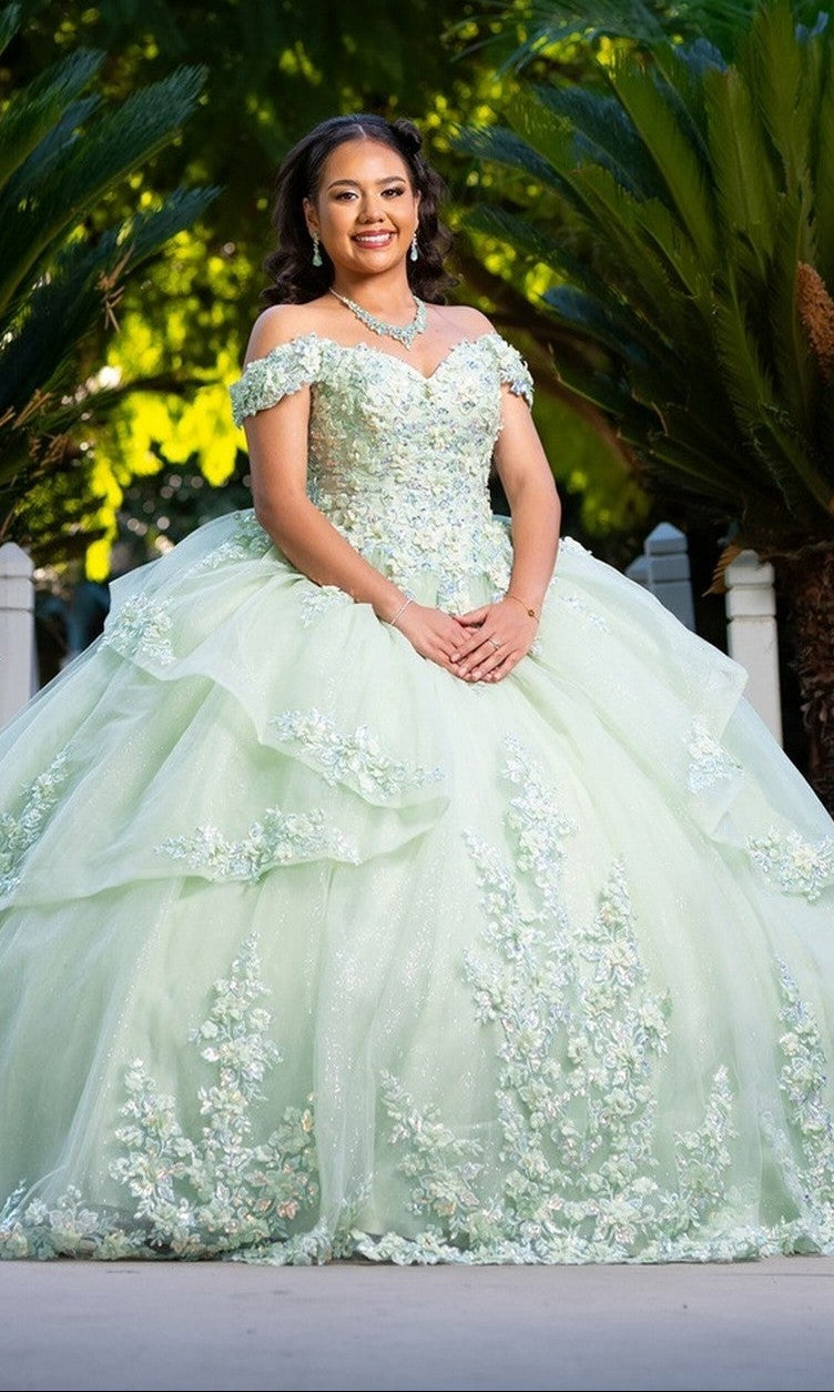 A young woman stands outdoors, smiling at the camera in the Quinceanera Dress CHF3104 By Chicas, featuring a sage color with a floral embroidered corset bodice and off-the-shoulder sleeves.