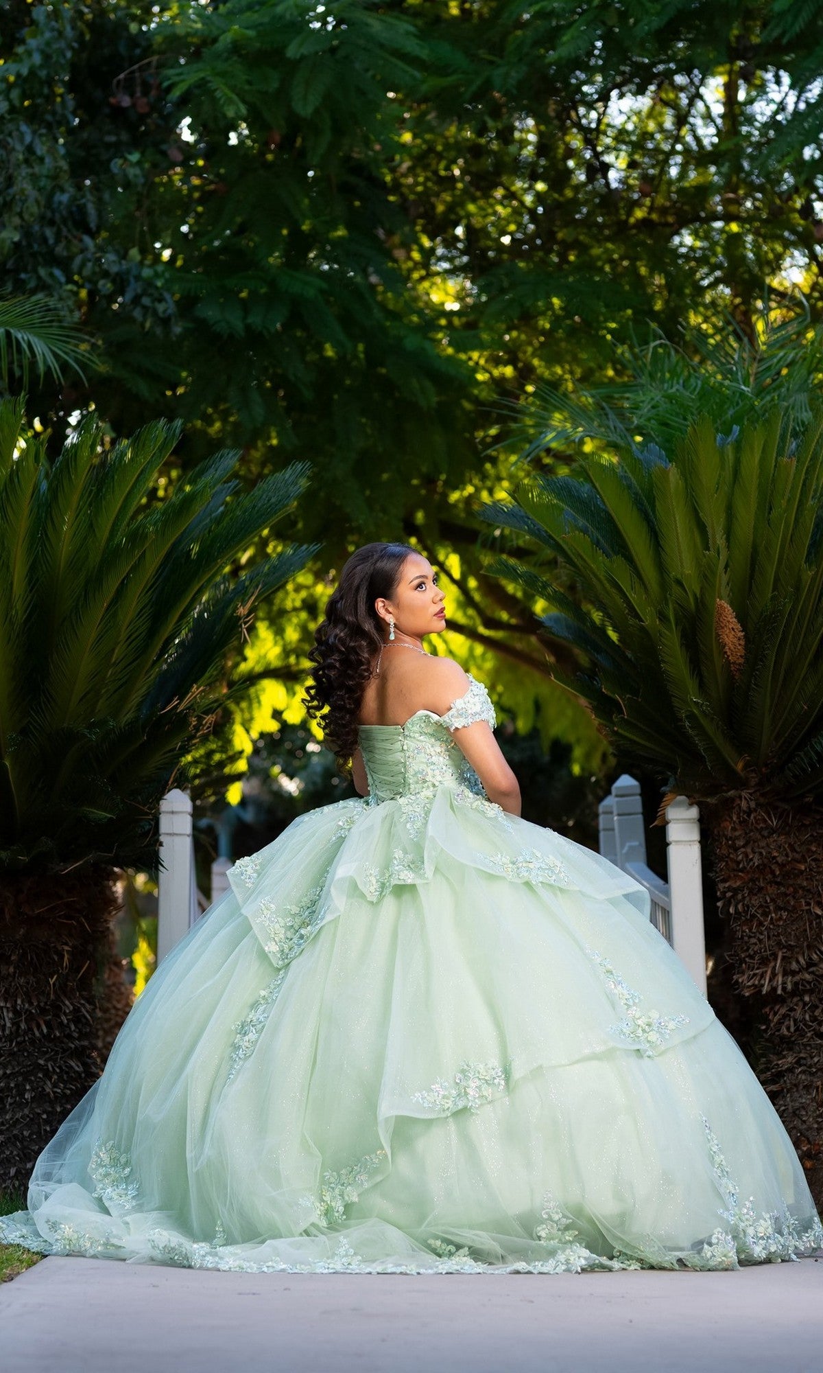 A young woman in a light green ball gown stands outdoors on a path, surrounded by lush green plants, looking over her shoulder. She wears the Quinceanera Dress CHF3104 By Chicas, which features a delicate floral embroidered corset bodice—an enchanting touch to this graceful scene.
