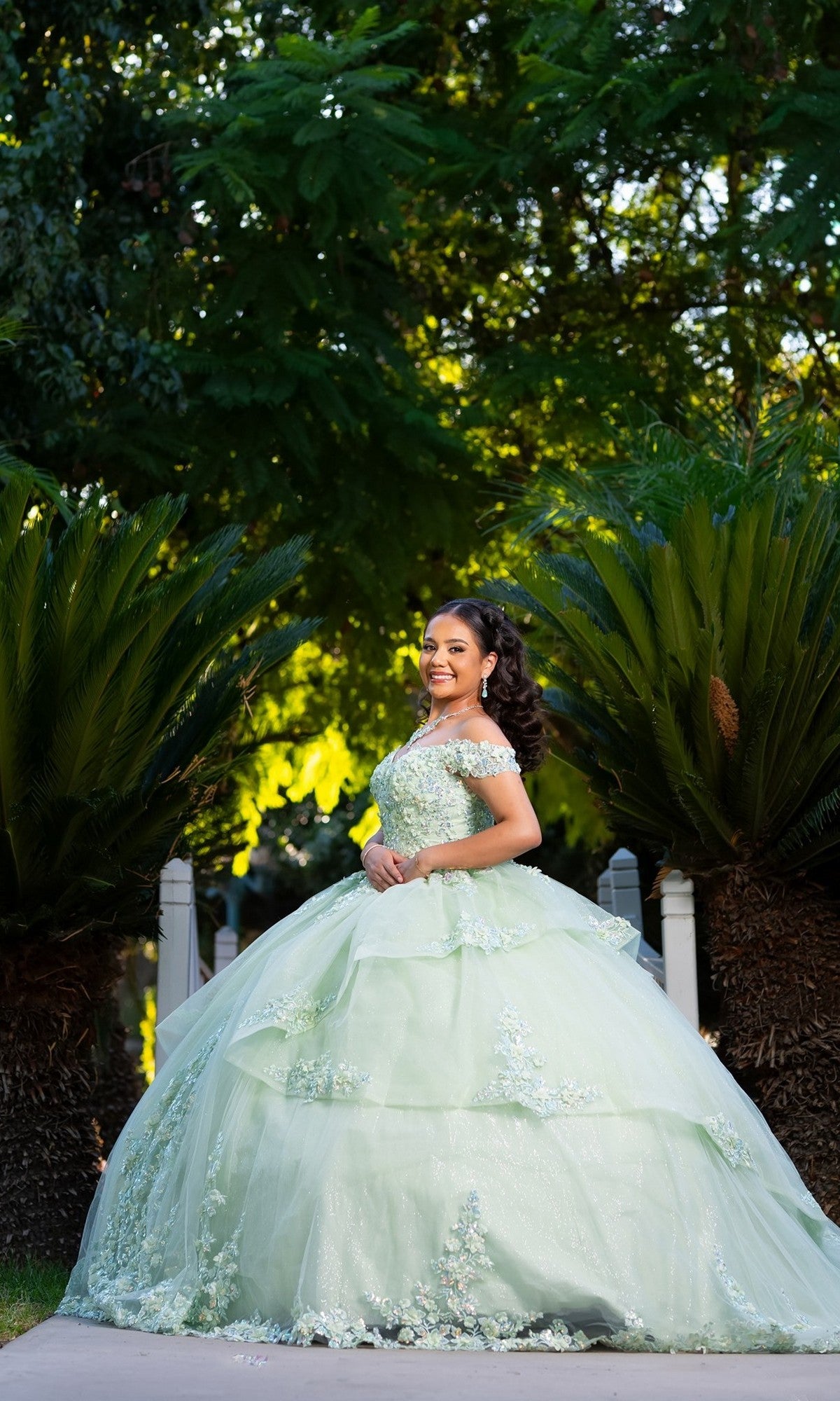 A young woman stands outdoors amidst lush greenery, smiling brightly in the “Quinceanera Dress CHF3104 By Chicas.” This sage green, off-the-shoulder gown features a beautifully floral embroidered corset bodice that adds an elegant touch to her enchanting look.