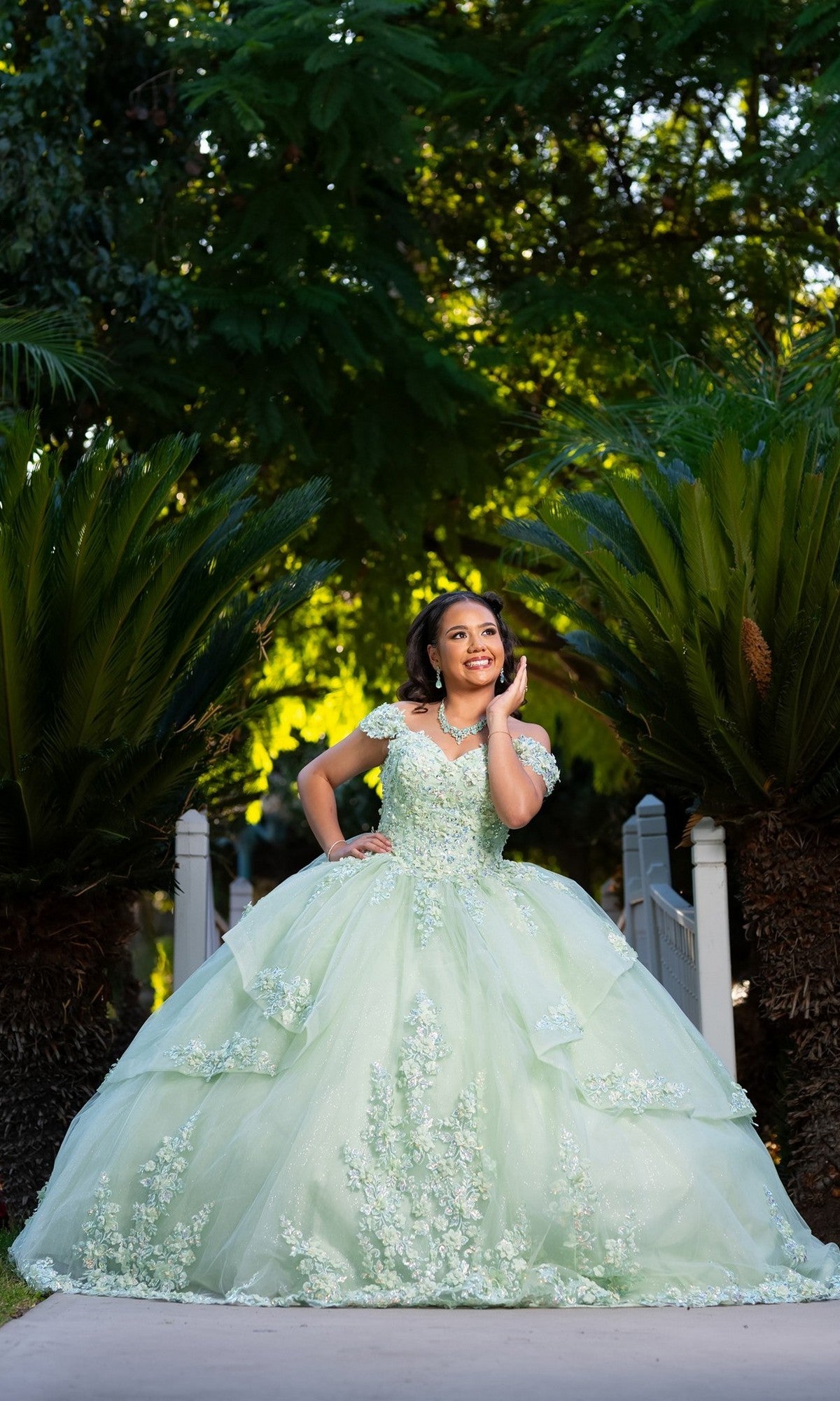 A young woman wearing the Quinceanera Dress CHF3104 By Chicas, a light green ball gown with a floral embroidered corset bodice, stands outdoors on a path between lush greenery, smiling and looking to the side with one hand near her face.