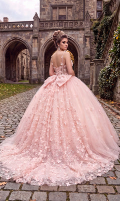 Wearing the Quinceanera Dress CHF3107 by Chicas—a pink ballgown featuring a corset bodice and delicate 3D floral appliques—a woman stands on a cobblestone path in front of a stone building with arches.