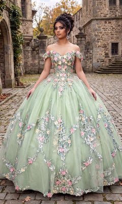 A woman stands on cobblestone in front of stone buildings, wearing the Quinceanera Dress CHF3109 By Chicas in sage—a pale green off-the-shoulder gown with a corset bodice and floral embellishments—in an outdoor setting with autumn trees.