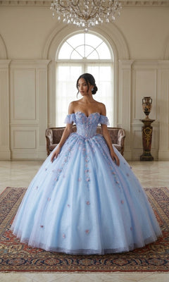 A woman stands indoors wearing the Quinceanera Dress CHF3114 By Chicas, a light blue, off-the-shoulder gown featuring delicate 3D floral appliques and a voluminous tulle skirt.